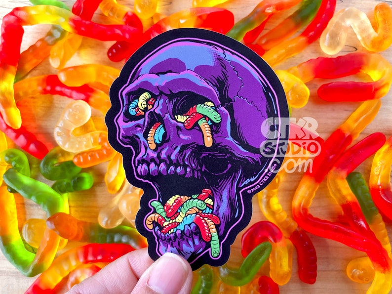 May include: A purple skull sticker with gummy worms coming out of its eyes and mouth. The sticker is on a background of gummy worms.
