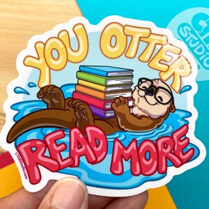 May include: A sticker featuring a cartoon otter wearing glasses and reading a stack of colorful books. The otter is floating in blue water with the text "You Otter Read More" in pink and yellow letters.