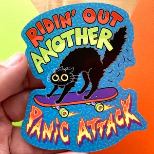 May include: A black cat riding a skateboard with the text "RIDIN' OUT ANOTHER PANIC ATTACK" in a colorful, retro style.