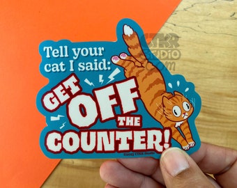 Puede incluir: Una pegatina azul y blanca con un gato naranja de dibujos animados saltando de una encimera. La pegatina dice "Dile a tu gato que dije: ¡BAJA DE LA ENCIMERA!" y tiene copyright de CTKR Studio.