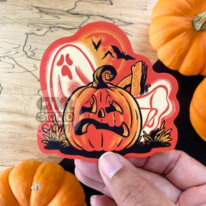May include: A vintage-style Halloween sticker featuring a jack-o'-lantern with a sad expression, a ghost, and a bat silhouette against an orange and red background. The sticker is held by a hand, with pumpkins in the background.