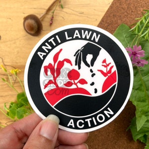 May include: A black and white sticker with the text "Anti Lawn Action" in a circle. The sticker features a hand sowing seeds into the ground with plants growing in the background. The sticker is on a wooden surface.