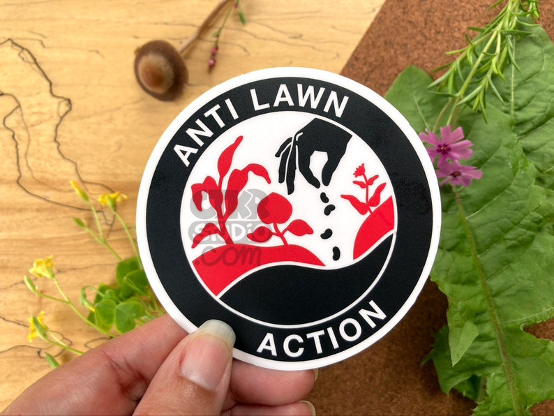 May include: A black and white sticker with the text "Anti Lawn Action" in a circle. The sticker features a hand sowing seeds into the ground with plants growing in the background. The sticker is on a wooden surface.
