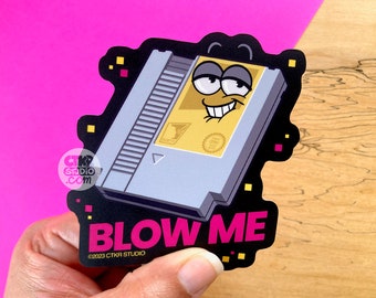 May include: A black and white sticker with a cartoon NES game cartridge with a face. The cartridge has a yellow face with a big smile and large eyes. The text "BLOW ME" is in pink letters on the sticker. The text "©2023 CTKR STUDIO" is in white letters below the pink text.