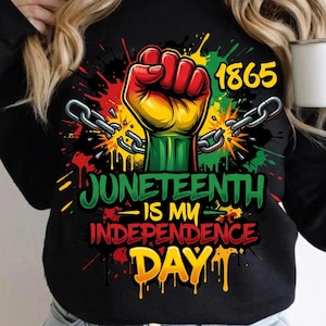 May include: Black long-sleeve shirt with a graphic celebrating Juneteenth. The design features a raised fist in red, yellow, and green, breaking chains. The text reads "Juneteenth is my Independence Day" with the year 1865.