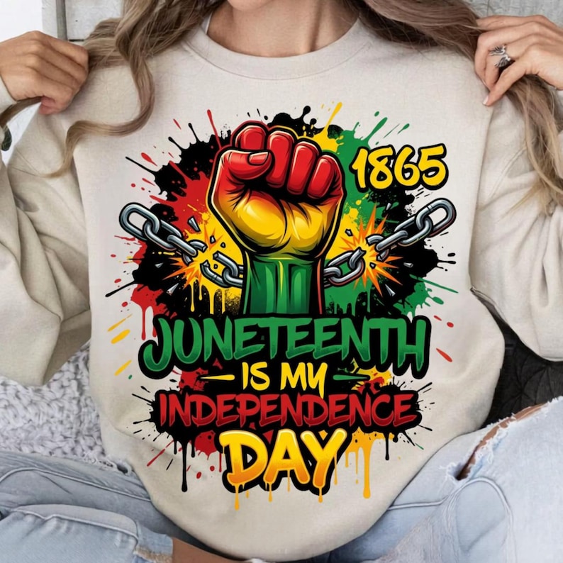 May include: A beige sweatshirt featuring a colorful graphic design. The design includes a raised fist in red, yellow, and green, chains, the number 1865, and the text "Juneteenth is my Independence Day."
