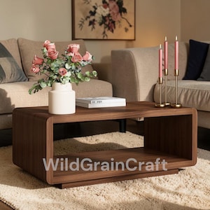 May include: A dark brown, modern coffee table with a rounded design. The table has a lower shelf and is placed on a cream-colored rug. A white vase with pink roses and a book sit on the table. The text "WildGrainCraft" is visible.
