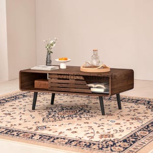 May include: A mid-century modern style coffee table made of dark wood with black legs. The table has a sliding door and a shelf for storage. A decorative rug with a floral pattern is beneath the table. The table is 48 inches long, 18 inches wide, and 16 inches high.