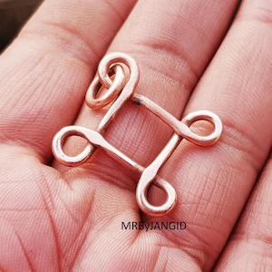 May include: A copper-colored metal hook and eye closure, held in a hand. The closure is square-shaped with looped ends. The text "MRByJANGID" is visible in the lower right corner.