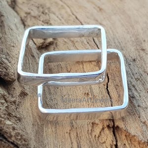 May include: Two interlocking square silver rings. The rings are made of a shiny metal and have a hammered texture. The rings are displayed on a wooden surface.