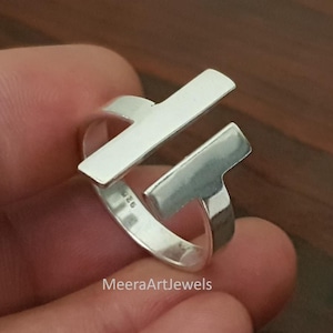 以下が含まれることがあります： モダンなデザインのシルバーリング。リングは、直角に交差する2つの長方形のバーが特徴です。リングは手で持たれています。「MeeraArtJewels」の文字が見えます。