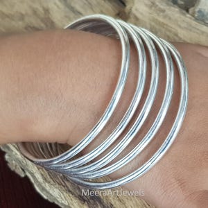 Puede incluir: Primer plano de seis pulseras plateadas apiladas en una muñeca. Las pulseras son lisas y redondas, reflejando la luz. El texto "MeeraArtJewels" es visible.