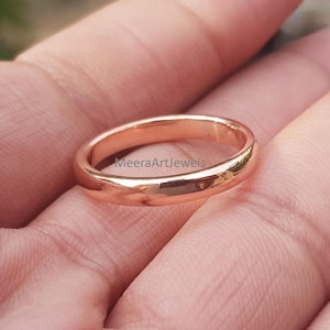 Puede incluir: Un anillo de banda de oro rosa pulido con una superficie lisa y redondeada. El metal tiene un tono rosa metálico cálido y un acabado reflectante. El texto MeeraArtJewels es visible en la superficie del anillo, indicando la marca de la joya.