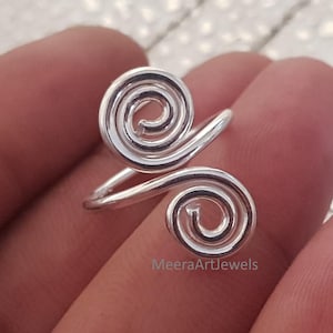 May include: A silver-colored ring with a double spiral design. The ring features two swirling, circular patterns connected by a band. The ring is shown against a blurred background.