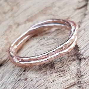 May include: A rose gold ring with a hammered texture. The ring is made of two intertwined bands. The ring is set against a wooden background. The text "MeeraArtJewels" is visible on the ring.