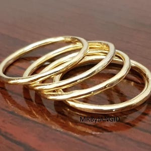 May include: Four gold-colored rings are stacked together, with a slightly hammered texture. The rings are on a dark brown wooden surface. The rings are close together, and the light reflects off the gold.
