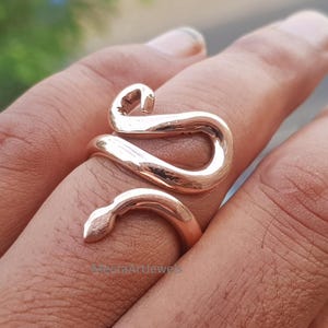 May include: A polished rose gold-toned metal ring shaped like a coiled snake that wraps around the finger. The design features a smooth, reflective surface with a tapered tail end and a distinct head shape. The text MeeraArtJewels is visible.