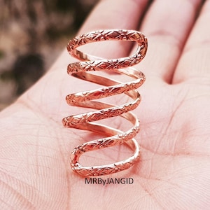 May include: A copper-colored, spiral ring with a textured, repeating floral pattern. The ring wraps around the finger multiple times, creating a unique and eye-catching design. The ring is shown on a hand. The text "MRByJANGID" is visible.