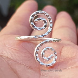 May include: A silver-colored bypass ring featuring two spiral ends with a hammered, textured finish. The metal surface reflects light, creating bright highlights across the curved, coiled design. The text MeeraArtJewels is visible at the bottom.