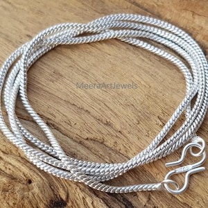 May include: A silver chain necklace with a woven texture, coiled on a wooden surface. The necklace has a hook clasp. The text "MeeraArtJewels" is visible on the wooden surface.