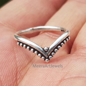 May include: A V-shaped silver ring featuring a beaded texture along the outer edge and a smooth inner band. The metal surface displays an oxidised finish with dark recessed areas. The stamp 925 is visible on the inner band of the ring.