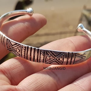 May include: A silver cuff bracelet with geometric patterns. The bracelet has a series of etched lines and shapes, including triangles and cross-like designs. The text "MRByJANGID" is visible on the bracelet.