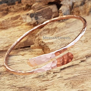 Peut inclure: Un bracelet manchette en cuivre martelé avec un design ouvert. Le bracelet a une surface texturée et une couleur brun-rougeâtre chaude. Les extrémités du bracelet sont légèrement évasées. Le texte "MeeraArtJewels" est visible.