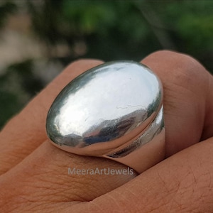 Peut inclure: Une bague en argent poli dotée d'un grand dôme ovale lisse. La surface métallique est très réfléchissante, révélant des reflets et des ombres subtils sur sa forme arrondie et organique. Le texte MeeraArtJewels est visible.