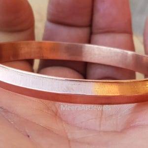 Puede incluir: Una pulsera de color cobre con un diseño plano y biselado. La joya es lisa y pulida, reflejando la luz. La pulsera mide aproximadamente 1,3 cm de ancho. El tema general es la joyería.