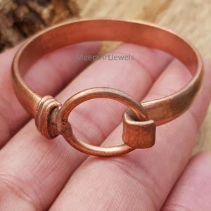May include: A copper-colored bracelet with an oval clasp. The bracelet is a solid band with a simple design. The clasp is a large oval that connects to the band. The bracelet is shown on a hand.