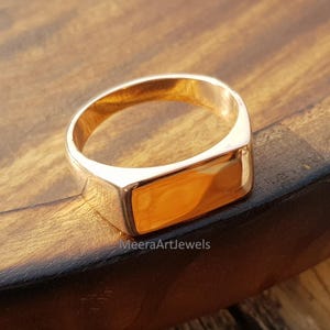 Puede incluir: Un anillo de sello dorado con una cara rectangular. El anillo está sobre una superficie de madera. El texto "MeeraArtJewels" es visible en la parte inferior derecha de la imagen.