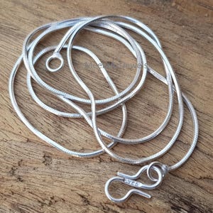 May include: A silver snake chain necklace with a round clasp and a small ring at the other end. The necklace is displayed on a wooden surface. The clasp is marked with the number "925".