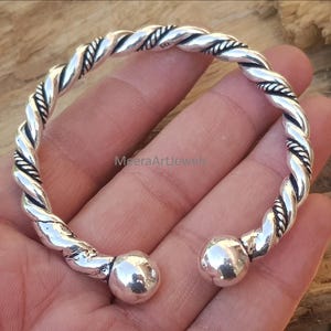 Peut inclure: Un bracelet manchette argenté avec un motif de corde torsadée. Le bracelet a une boule arrondie à chaque extrémité. Le bracelet est présenté sur une main.