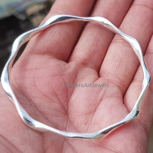 Puede incluir: Un brazalete de plata pulida con un diseño de borde ondulado y orgánico continuo. La banda metálica circular tiene una superficie lisa y reflectante, con una silueta ondulante distintiva que crea una serie de picos y valles suaves.