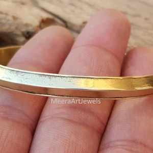 Puede incluir: Un brazalete de color dorado con una superficie plana y ligeramente texturizada. El brazalete tiene un diseño simple y elegante y probablemente está hecho de latón o un metal similar. El brazalete se muestra en una mano.