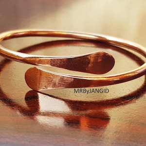 May include: A copper-colored cuff bracelet with an open design. The bracelet has a smooth, rounded band that curves around the wrist, with flattened, rounded ends that overlap. The surface reflects light, creating highlights and shadows. The text "MRByJANGID" is visible.