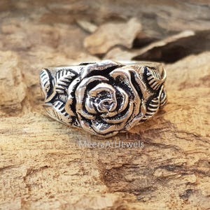 May include: A silver-colored ring featuring a detailed rose design. The ring showcases a prominent rose in full bloom, surrounded by leaves. The text "MeeraArtJewels" is visible on the surface. The ring is set against a textured, brown background.