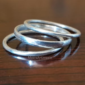 Op de afbeelding: Drie zilveren ringen gestapeld op een donkerbruin oppervlak. De ringen zijn eenvoudige banden met een licht getextureerde uitstraling. De ringen liggen dicht bij elkaar, waardoor een gelaagd effect ontstaat. De ringen zijn reflecterend.