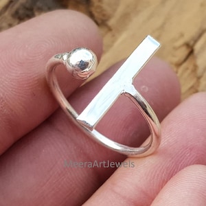 以下が含まれることがあります： モダンなデザインのシルバーリング。リングは、小さなテクスチャードボールとストレートバー要素が特徴です。リングは木製の表面に表示されています。リングはジュエリーです。