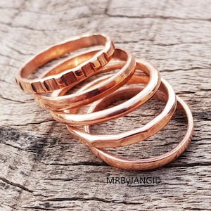Puede incluir: Un conjunto de anillos de cobre con diferentes texturas. Algunos anillos tienen un acabado liso y pulido, mientras que otros presentan un diseño martillado o texturizado. Los anillos se muestran sobre una superficie de madera rústica.
