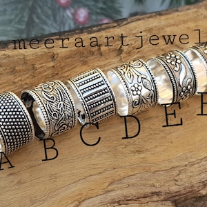 以下が含まれることがあります： さまざまなエンボス加工のデザインが施されたシルバーリングのコレクション。ドット、花柄、幾何学模様など。リングは木製の表面に展示されており、上部には「meeraartjewels」の文字が見えます。