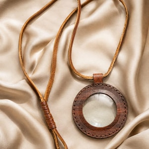 Può includere: Un ciondolo a forma di lente d'ingrandimento circolare con una cornice in legno marrone scuro e cuciture visibili lungo il bordo. La lente è trasparente e inserita nel legno. Un lungo cordino in pelle marrone è fissato alla sommità.