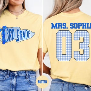 May include: A pale yellow t-shirt featuring a blue and white gingham pennant graphic on the front with the text 3RD GRADE and a blue bow. The back displays the text MRS. SOPHIA above the large blue and white gingham numbers 03 in a varsity font.