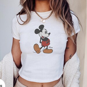 Puede incluir: Top corto blanco con un gr&aacute;fico de Mickey Mouse. El personaje de dibujos animados est&aacute; representado en negro, rojo y marr&oacute;n. El logotipo de Disney es visible. El top es de manga corta.