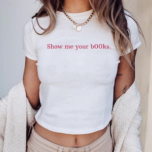 May include: White crop top with the text "Show me your books" in pink. The shirt is short-sleeved and has a round neckline. A gold chain necklace is visible. The person is wearing beige pants and a cream-colored cardigan.