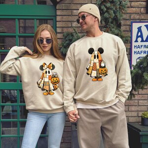 May include: Two beige crewneck sweatshirts featuring a graphic of a ghost wearing Mickey Mouse ears and a patchwork-style cloak. The ghost holds a jack-o'-lantern bucket. The left sweatshirt graphic includes a bow, while the right one does not.