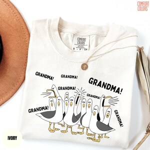May include: Ivory-colored t-shirt with the word "GRANDMA!" repeated around a cartoon illustration of seagulls. The shirt has a Comfort Colors tag and a gold necklace is draped across it.
