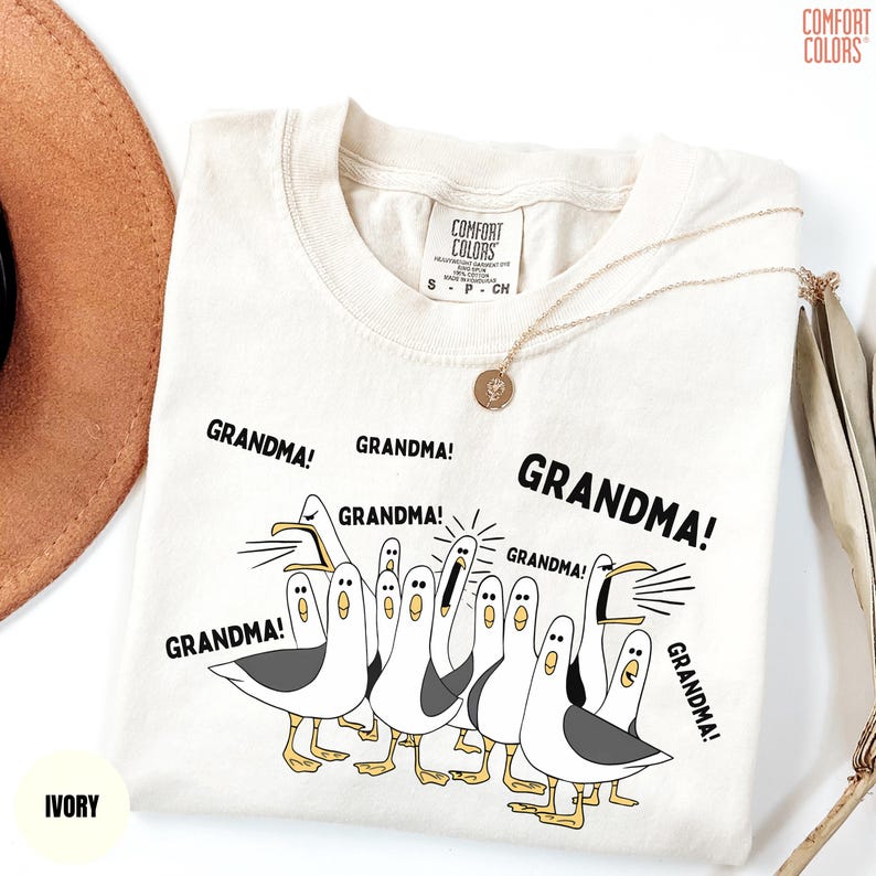 May include: Ivory-colored t-shirt with the word "GRANDMA!" repeated around a cartoon illustration of seagulls. The shirt has a Comfort Colors tag and a gold necklace is draped across it.