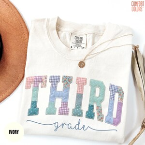 May include: Ivory-colored t-shirt with the word "THIRD" in patchwork letters, and the word "grade" in a cursive font. A gold necklace with a pendant rests on the shirt. The shirt is from Comfort Colors.