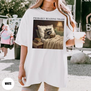 以下が含まれることがあります： 「PROBABLY READING TRASH」の文字がプリントされた白いTシャツ。その上には、ベッドで本を読むアライグマの額縁入り画像。画像は、ランプと枕のある居心地の良い環境にいるアライグマを描いています。Tシャツはゆったりとしたオーバーサイズフィットです。